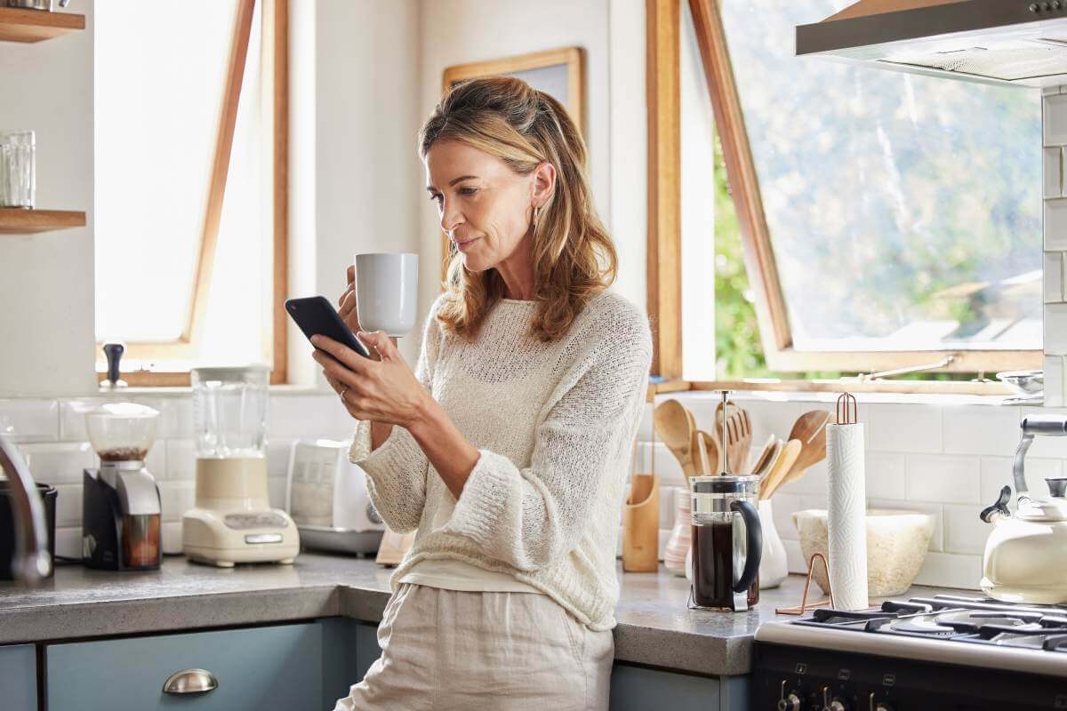 Mulher loira de meia-idade encostada na bancada de uma cozinha. Ela toma café enquanto olha o celular. 