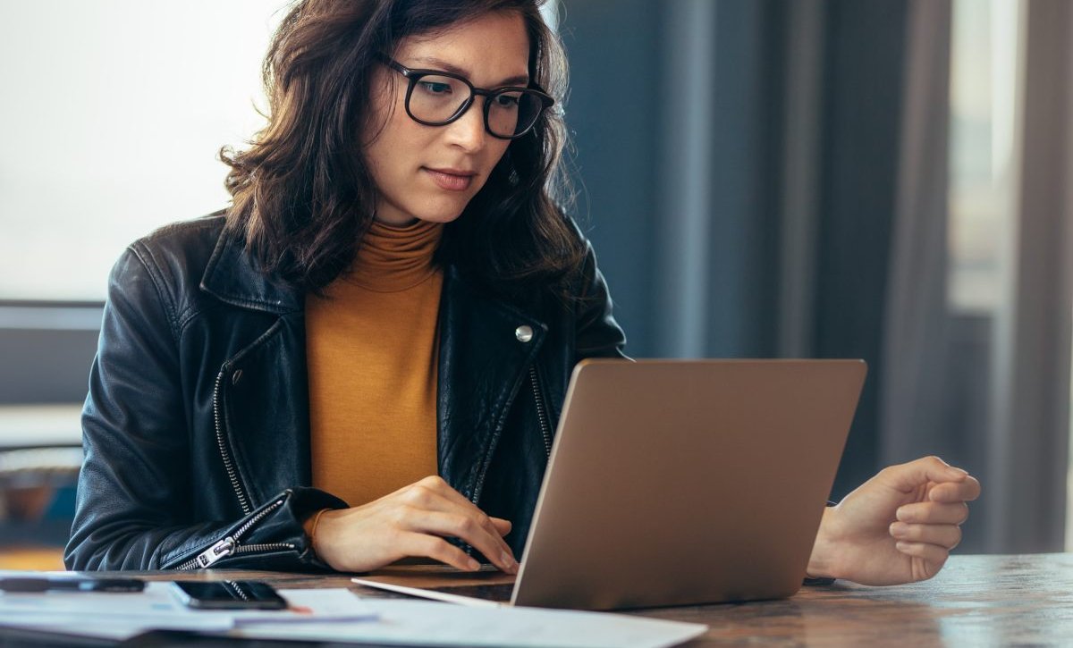 Mulher em ambiente de escritório digitando em um computador com a mão direita. Ela usa óculos, uma blusa cor mostarda e uma jaqueta de couro.