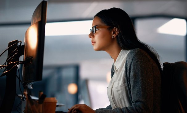 Mulher de óculos digitando em teclado de desktop. A tela não é visível. A mulher está em um escritório, veste camisa branca e casaco azul.