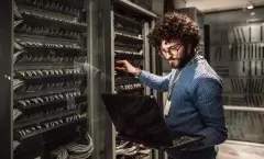 Homem moreno de barba e camisa azul segurando um notebook enquanto faz configurações em um servidor. Ele aparentemente está em uma empresa e usa o notebook para checar algum site para realizar as configurações.