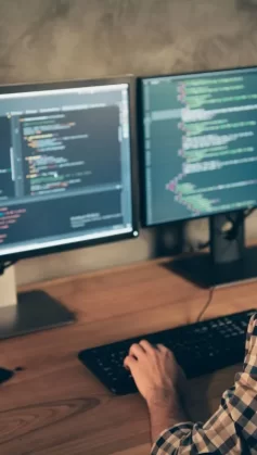 Homem de cabelo escuro, barba, óculos e camisa xadrez trabalhando em dois desktops ao mesmo tempo. Ele está de costas para a câmera. Nas telas, aparecem códigos de programação.