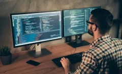Homem de cabelo escuro, barba, óculos e camisa xadrez trabalhando em dois desktops ao mesmo tempo. Ele está de costas para a câmera. Nas telas, aparecem códigos de programação.