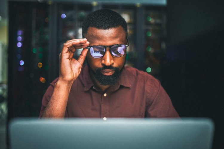 Homem negro de óculos e camisa marsala olhando para um computador enquanto analisa códigos de programação. Ele segura os óculos com a mão direita. Atrás dele parece haver um servidor.