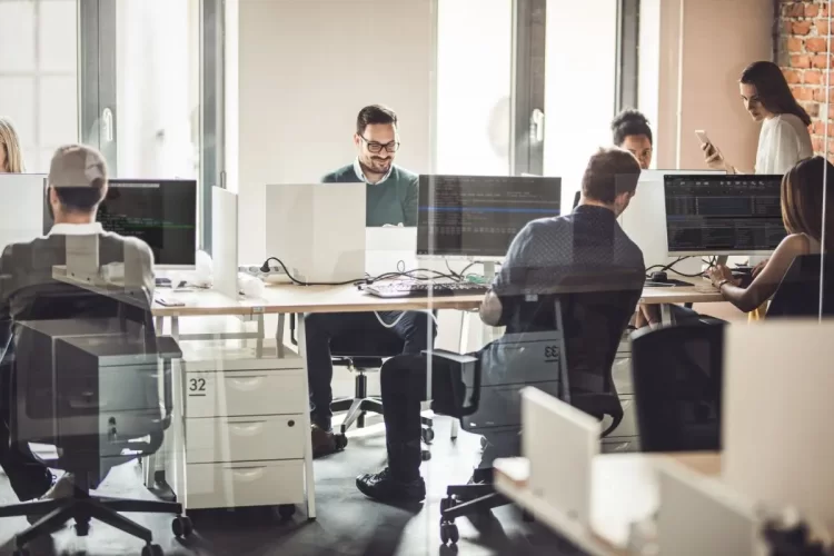 Grupo de pessoas programadoras trabalhando em um escritório e conversando sobre programação. Eles estão todos sentados ao redor de uma grande mesa.