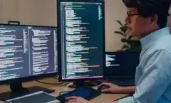 Homem de traços asiáticos, usando óculos e camisa, trabalha com duas telas de computador. Nelas, podemos ver códigos de uma linguagem de programação não identificada.