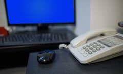 Um telefone branco sobre uma mesa de escritório. Há um mouse preto perto dele e um teclado com uma tela azul ao fundo. Possivelmente, são equipamentos de um escritório.
