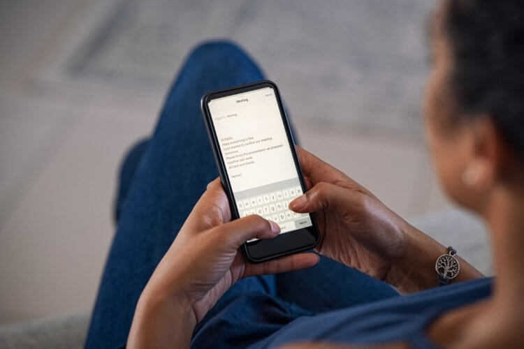 A imagem mostra uma mulher negra usando camiseta azul e calça jeans azul enquanto segura um smartphone: Ela está digitando um e-mail, que será enviado para um de seus contatos profissionais.