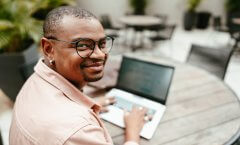 Um homem negro de óculos usando camisa social está digitando informações em um notebook. Ele aparentemente está em um café durante o dia, e há outras mesas ao fundo para complementar o ambiente.