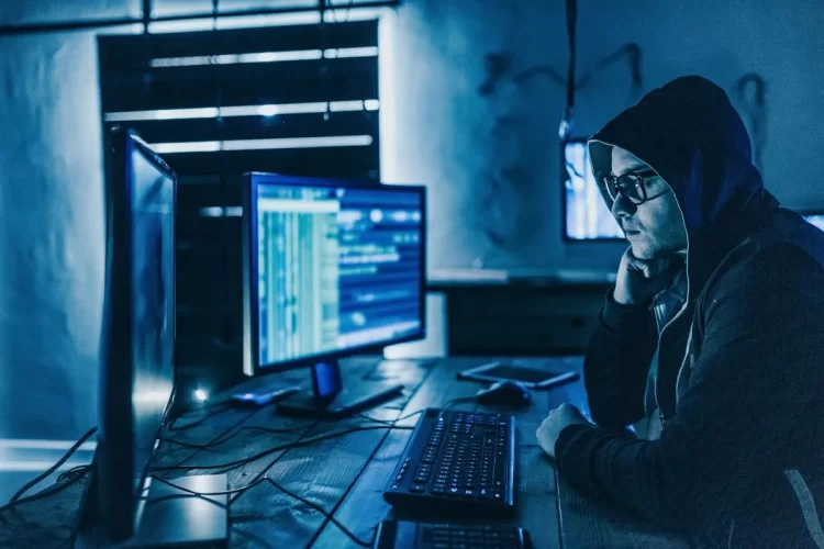 A imagem mostra um homem pensativo enquanto observa uma das telas de seu computador. Ele está usando óculos e um capuz para cobrir a sua cabeça.