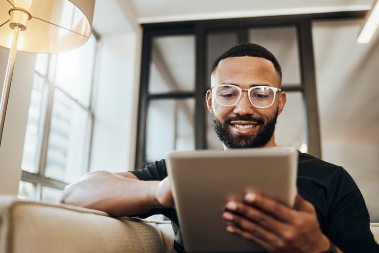 A imagem mostra um homem negro na sala de sua casa mexendo em um tablet. Ele possivelmente está acessando algum site que conta com recursos de CDN, o que melhora a sua experiência de navegação.