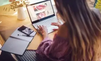 A imagem mostra uma mulher de costas para a câmera, trabalhando em um notebook. Ela está editando um blog de culinária e, para isso, precisa de um CMS. Ao redor dela, sobre a mesa, há um caderno aberto, uma caneta e um celular. Atrás do computador temos um vaso de flores.