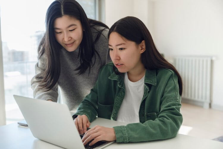 A imagem mostra duas mulheres asiáticas de idades diferentes. Uma delas está mexendo no notebook, enquanto a outra aparece para avaliar o que está na tela. Elas aparentemente estão em casa, pois há um sistema de aquecimento para os dias frios ao fundo.