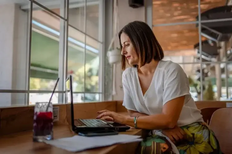 A imagem mostra uma mulher de blusa branca e cabelos curtos mexendo em um notebook. Há grandes chances de que ela esteja em um escritório, já que há outras cadeiras por perto.