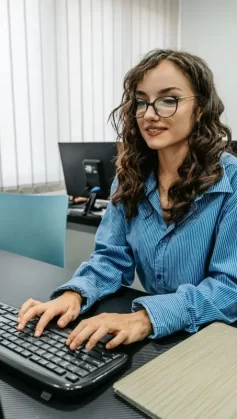 A imagem mostra uma mulher de óculos sentada de frente para um computador. Ela está com uma camisa azul e digita algo no teclado. Há outra pessoa na cadeira ao fundo, completando o cenário.