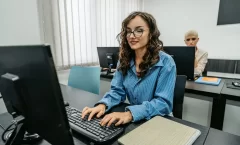 A imagem mostra uma mulher de óculos sentada de frente para um computador. Ela está com uma camisa azul e digita algo no teclado. Há outra pessoa na cadeira ao fundo, completando o cenário.