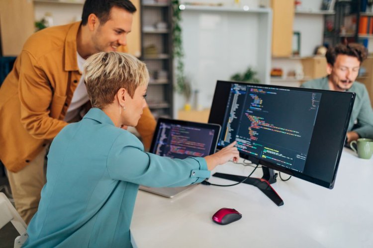 A imagem mostra um homem e uma mulher conversando sobre programação. A mulher aparentemente está cuidando de escrever o código, e mostra para o homem o andamento da codificação, apontando alguns elementos que aparecem no monitor.