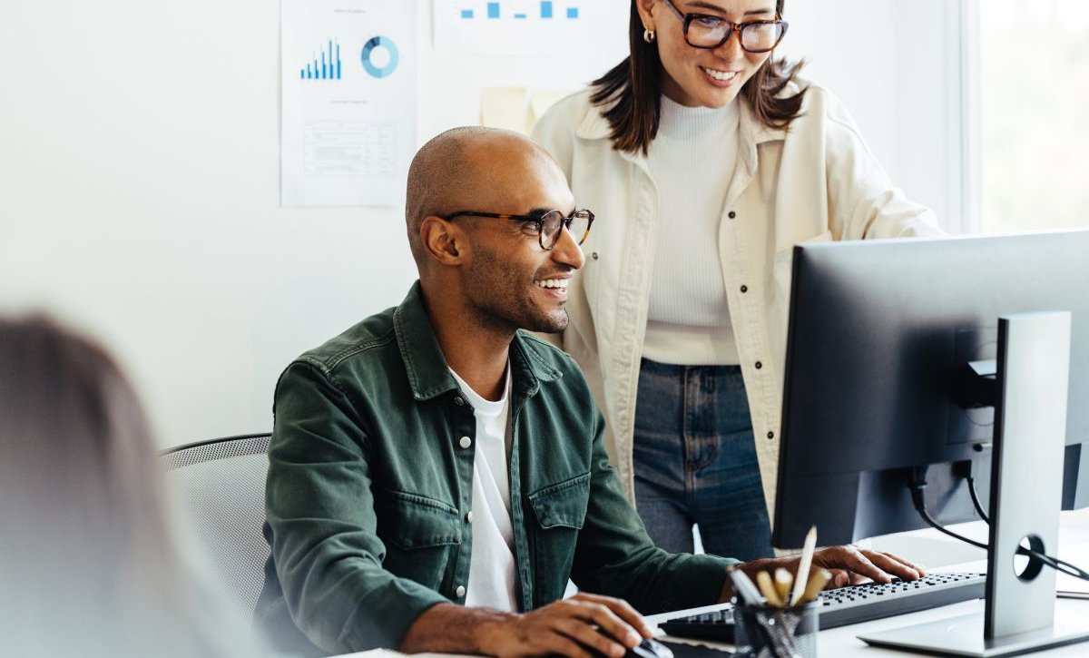 A imagem mostra um homem e uma mulher conversando em um escritório. Eles vestem blusas e usam óculos, e estão falando sobre o desenvolvimento de um layout de site. Outra pessoa aparece desfocada na imagem, mas ela não participa da conversa.