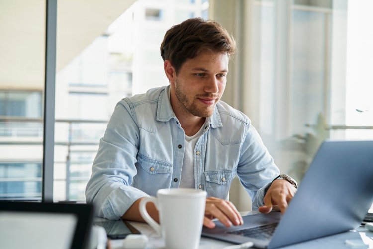 A imagem mostra um homem branco trabalhando em um escritório. Ele está lidando com um layout de site, por isso mexe no notebook em uma área reservada, onde há apenas ele. Há uma caneca de café do lado dele.