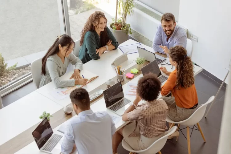 A imagem apresenta um grupo multiétnico de pessoas sentadas ao redor de uma mesa, com notebooks sobre ela, em uma sala de reuniões. Elas conversam e sorriem entre si. Vestem roupas casuais. O tema do artigo é lead scoring.