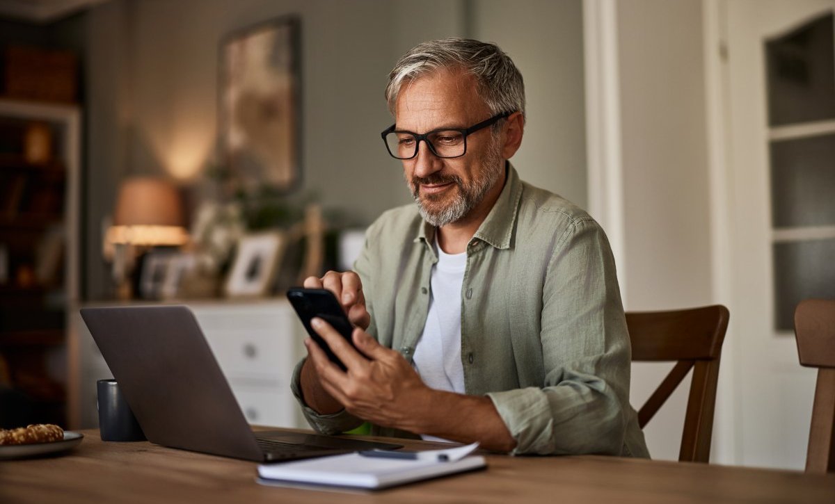 A imagem mostra um homem de meia-idade diante de um notebook e com um celular na mão. Após alguns trabalhos com localhost, ele verifica o funcionamento de uma aplicação no smartphone.