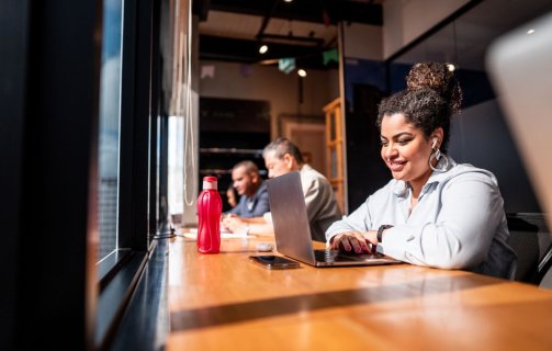 A imagem mostra uma mulher em um escritório diante de uma janela, trabalhando em otimização de sites. Ela está tocando o touchpad do notebook. Ao fundo, é possível ver mais três pessoas trabalhando no mesmo ambiente.