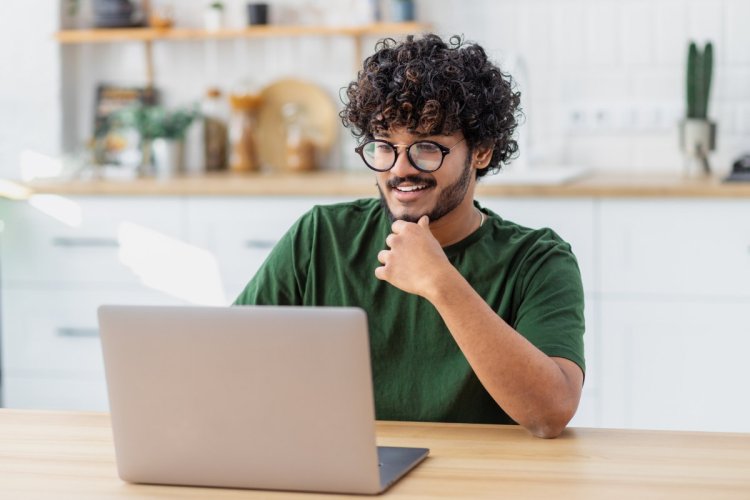 A imagem mostra um homem trabalhando sentado em sua casa. Ele está vestido com roupas casuais, digitando no teclado do notebook, e há alguns objetos em cima de um balcão ao fundo.