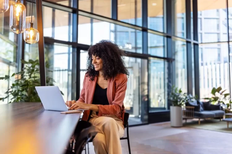 A imagem mostra uma mulher de roupa social em um café. Ela está olhando para a tela do notebook enquanto trabalha com a otimização de sites. Há outras cadeiras no ambiente, mas elas estão vazias.