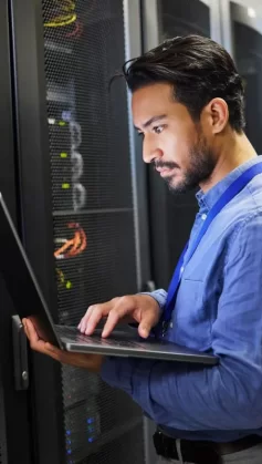 A imagem mostra um homem de barba vestindo uma camisa social azul e segurando um notebook. Ele está analisando a infraestrutura de servidor web de uma empresa, em uma sala com diversos servidores instalados.