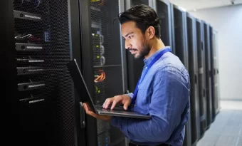 A imagem mostra um homem de barba vestindo uma camisa social azul e segurando um notebook. Ele está analisando a infraestrutura de servidor web de uma empresa, em uma sala com diversos servidores instalados.