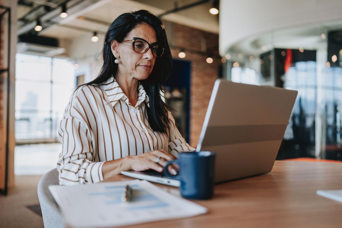 A imagem mostra uma mulher de meia-idade usando roupa social em um escritório. Ela está diante de um notebook, trabalhando na construção de sites e otimizando imagens WebP para deixá-los mais rápidos.