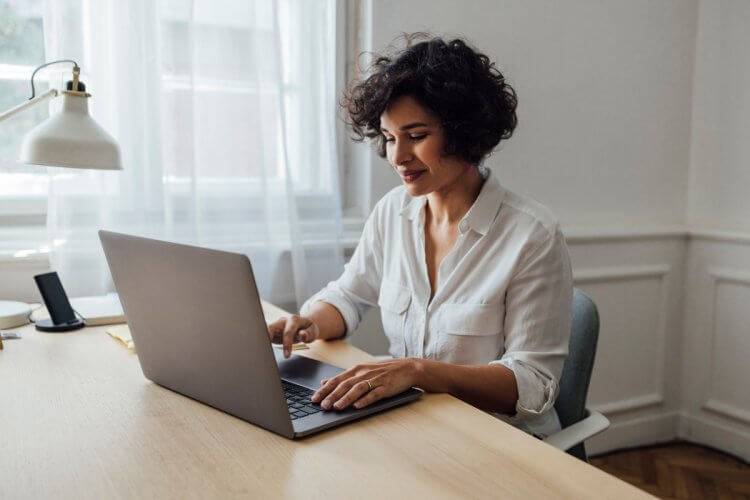 A imagem mostra uma mulher jovem digitando no teclado de um notebook. Ela está sozinha no que parece ser um pequeno escritório em casa, e está procurando por um bom cliente FTP para realizar transferências de arquivos para outras pessoas.