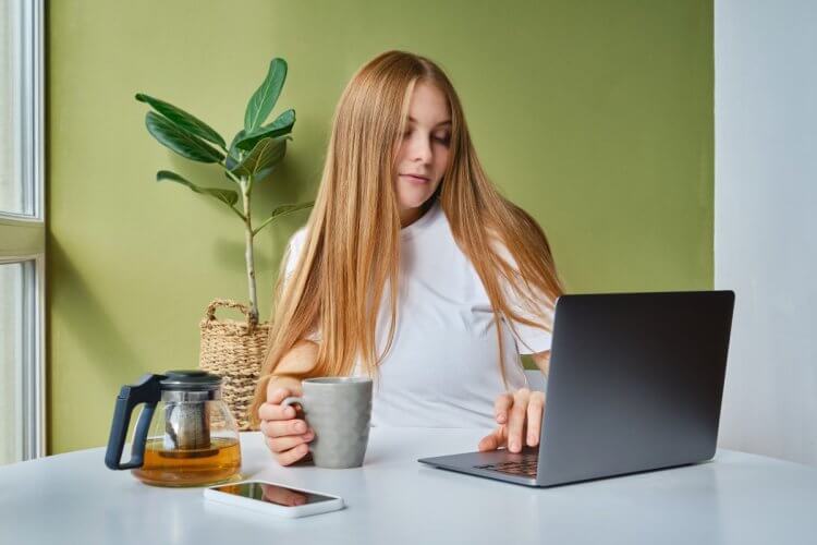 A imagem mostra uma mulher tomando uma xícara de café enquanto faz pesquisas em um notebook. Ela está avaliando as melhores opções de cliente FTP existentes no mercado, e faz isso no que parece ser um escritório.