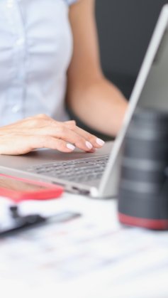 A imagem mostra uma mulher branca sentada com um notebook à sua frente. O rosto dela não aparece. Há uma câmera e uma lente em plano desfocado e ela possivelmente está usando uma IA que cria vídeos para complementar o seu trabalho.