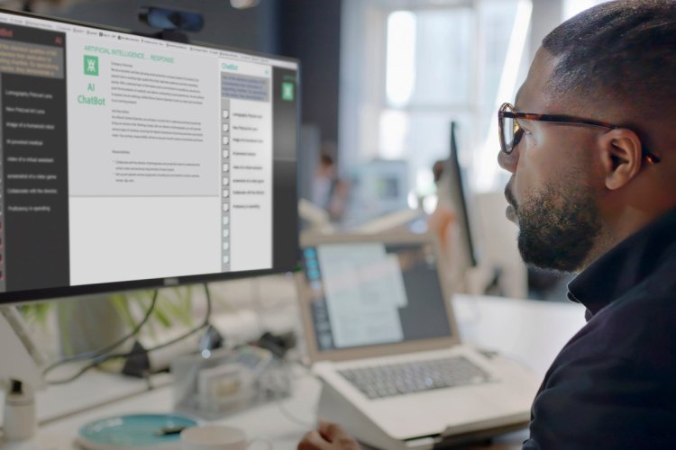 A imagem mostra um homem negro de óculos, diante de um computador, utilizando uma Inteligência Artificial para criar textos grátis. Ele está em um escritório, e há outras mesas e computadores na imagem para complementar o ambiente. 