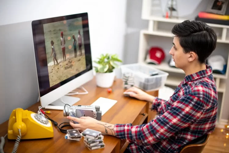 A imagem mostra um rapaz editando um vídeo em um computador. A gravação no monitor mostra algumas pessoas. Ele aparentemente está fazendo este trabalho em home office, já que há uma estante ao fundo, um vaso de plantas e um telefone antigo.