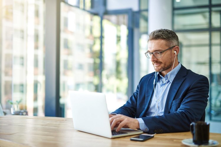 A imagem mostra um homem de roupa social e óculos trabalhando em um escritório. Ele está mexendo em um notebook, no qual faz a análise de gráficos gerados por Inteligência Artificial enquanto ouve música em um fone de ouvido.