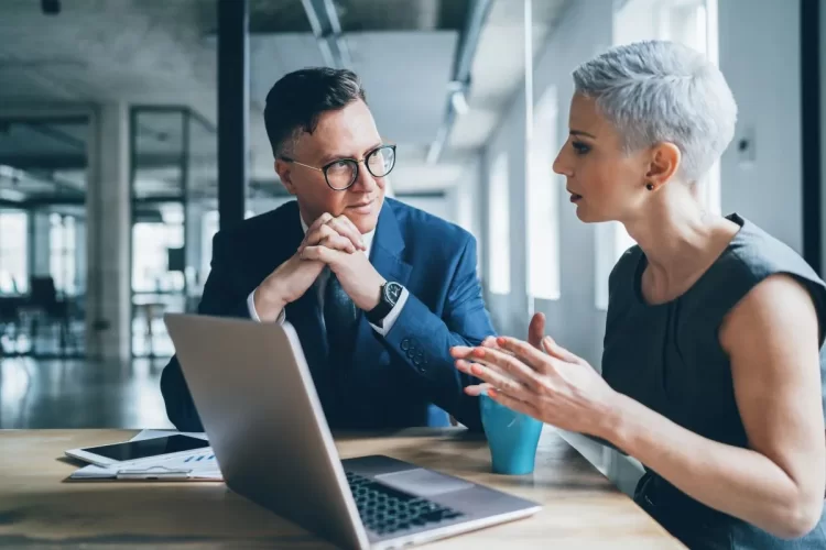 A imagem mostra um homem e uma mulher conversando em um escritório. Eles estão sentados diante de um notebook, no qual visualizam informações sobre sites de Inteligência Artificial e decidem quais ferramentas vão utilizar para as atividades da empresa.