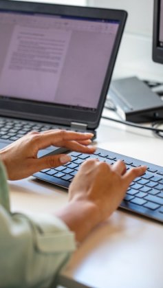 A imagem mostra uma mulher digitando em um teclado de computador em uma mesa de escritório. O rosto dela não aparece. Ela tem um notebook com um documento aberto ao lado, no qual checa um texto que foi reescrito com a Inteligência Artificial do ChatGPT.