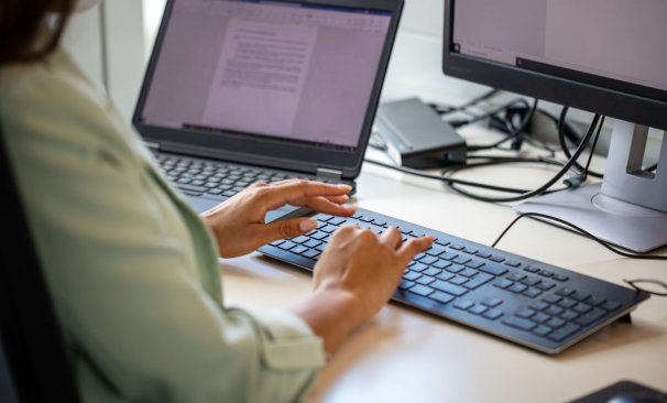 A imagem mostra uma mulher digitando em um teclado de computador em uma mesa de escritório. O rosto dela não aparece. Ela tem um notebook com um documento aberto ao lado, no qual checa um texto que foi reescrito com a Inteligência Artificial do ChatGPT.