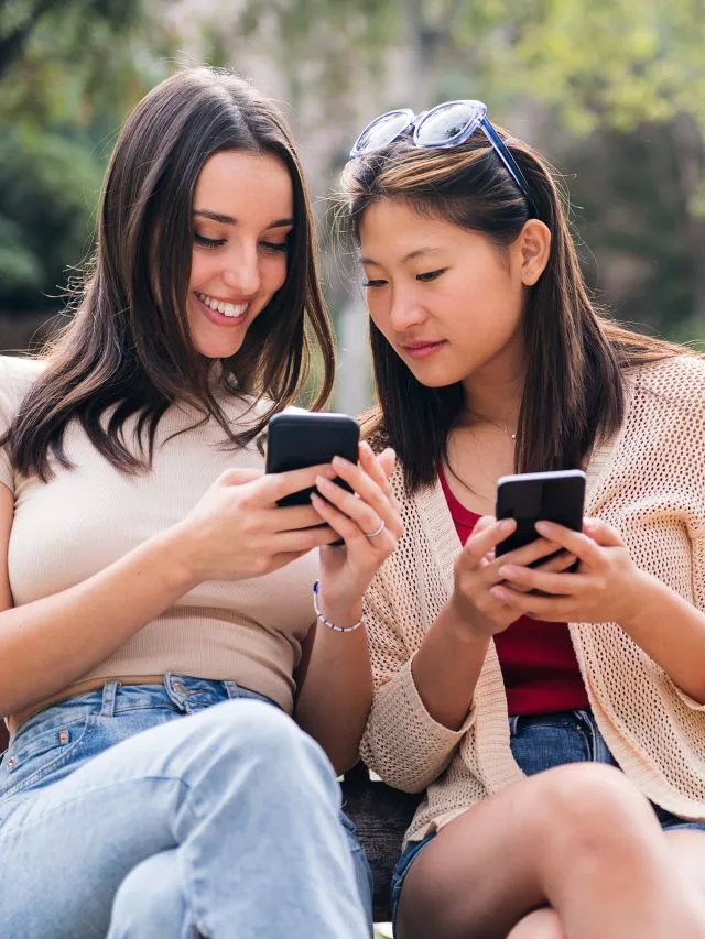 Duas jovens mulheres, uma branca e uma asiática vestindo roupas casuais estão sentadas usando os seus smartphones. Elas parecem estar em uma área externa, com a presença de plantas ao fundo.