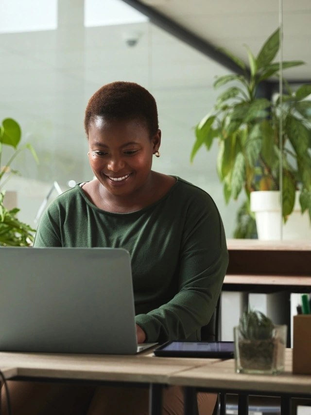 Uma mulher jovem e afro-americana, de cabelos curtos e blusa de manga comprida verde-escuro, está em um escritório decorado com vasos de plantas, usando seu notebook para pesquisar algo na internet.