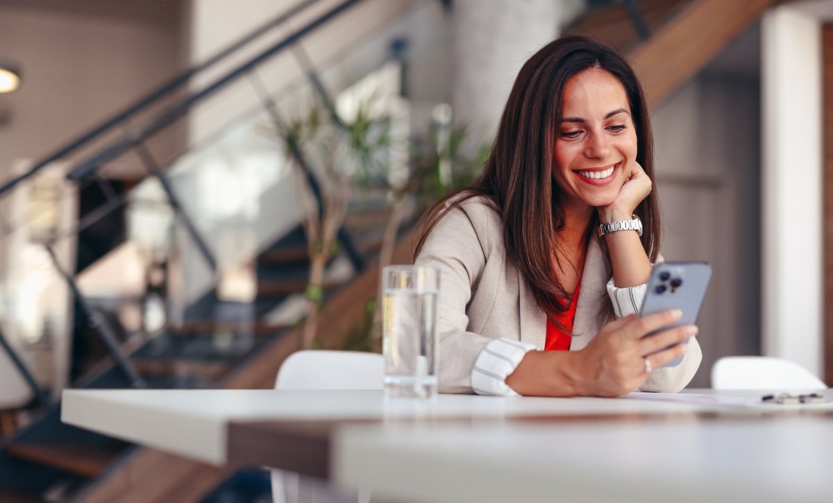 A imagem mostra uma mulher em uma casa usando um celular. Ela está apoiada em uma mesa com um copo de água à frente e olha para o smartphone após ter sido impactada por alguma publicação que fez uso de impulsionamento com a ajuda do TikTok Ads.