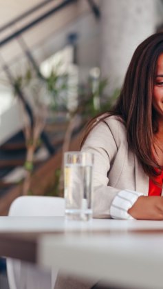 A imagem mostra uma mulher em uma casa usando um celular. Ela está apoiada em uma mesa com um copo de água à frente e olha para o smartphone após ter sido impactada por alguma publicação que fez uso de impulsionamento com a ajuda do TikTok Ads.