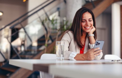 A imagem mostra uma mulher em uma casa usando um celular. Ela está apoiada em uma mesa com um copo de água à frente e olha para o smartphone após ter sido impactada por alguma publicação que fez uso de impulsionamento com a ajuda do TikTok Ads.