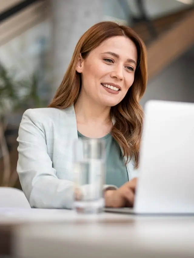 Uma mulher jovem usando roupas casuais está em frente a uma mesa de escritório com um notebook, digitando um texto.