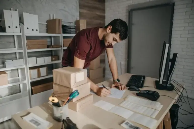 A imagem mostra um homem em um armazém anotando algo em uma folha de papel. Ele é dono de um comércio online que possivelmente trabalha com produtos para revenda, já que há dois pacotes sobre a mesa e caixas variadas na prateleira que está ao fundo do ambiente. 