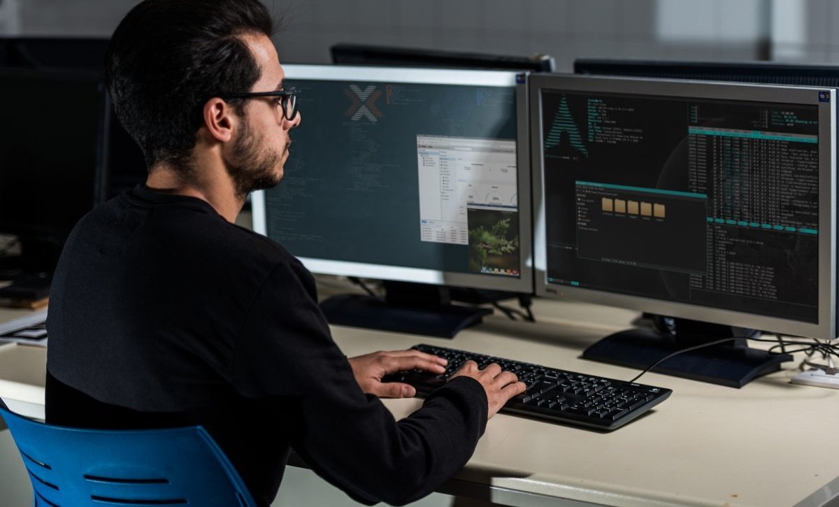 A imagem mostra um homem jovem em frente à mesa de trabalho dele. Ele está digitando no teclado enquanto verifica linhas de código geradas com o auxílio do SCP Linux.