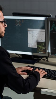 A imagem mostra um homem jovem em frente à mesa de trabalho dele. Ele está digitando no teclado enquanto verifica linhas de código geradas com o auxílio do SCP Linux.