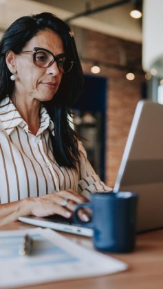 A imagem mostra uma mulher trabalhando em um escritório. Ela está digitando em um notebook o endereço de um site confiável, checando com cuidado todos os elementos para não cair em nenhum tipo de golpe.