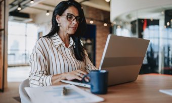 A imagem mostra uma mulher trabalhando em um escritório. Ela está digitando em um notebook o endereço de um site confiável, checando com cuidado todos os elementos para não cair em nenhum tipo de golpe.